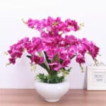 Sur un meuble, on voit un pot de fleurs blanc avec des orchidées artificielles violettes à l'intérieur.