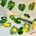 Guirlande lumineuse allumée avec feuilles artificielles de Monstera à moitié suspendue et posée sur une surface blanche avec des livres posés dessus