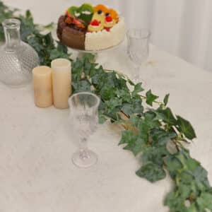 Branche de lierre posée sur une table avec une nappe blanche, des verres à pieds, des bougies et un gâteau aux fruits.