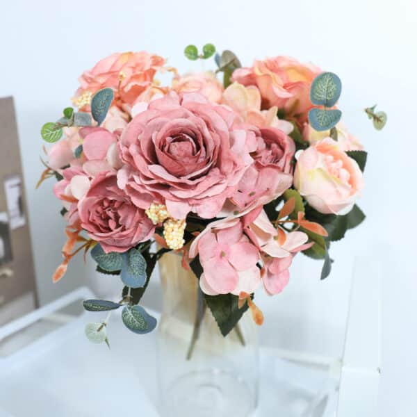 Un bouquet de roses et de pivoines au style vintage dans un vase transparent sur une commande et devant un mur blanc.