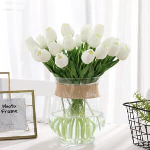 Bouquet de tulipes blanches dans une vase transparent posé sur une table avec une nappe blanche, un cadre et un rideau blanc derrière