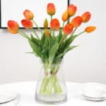 Bouquet de tulipes oranges dans un vase transparent posé sur une table dressée avec des assiettes et une tasse.