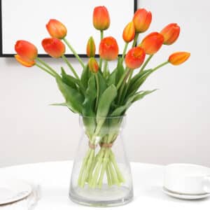 Bouquet de tulipes oranges dans un vase transparent posé sur une table dressée avec des assiettes et une tasse.