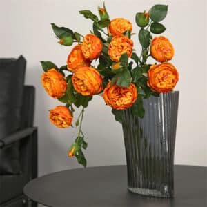 Bouquet de rose orange avec des feuilles verte dans un vase transparent gris sur une table grise sur un fond beige.