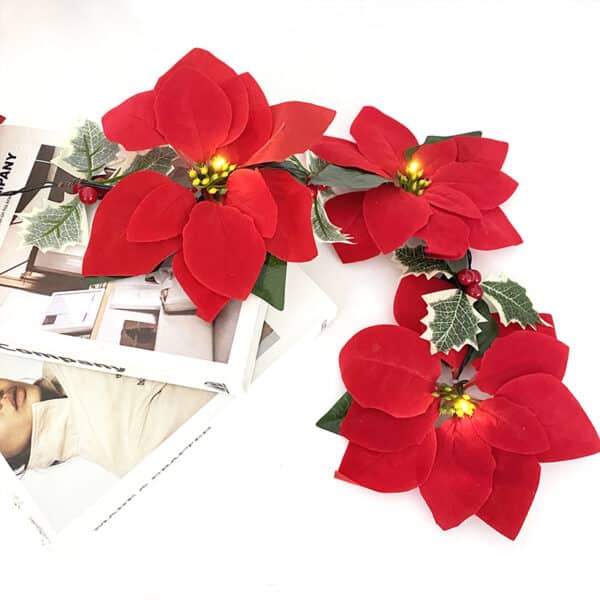 Guirlande avec fleurs rouges artificielles de Noël avec du houx posée sur un fond blanc sur des magazines.