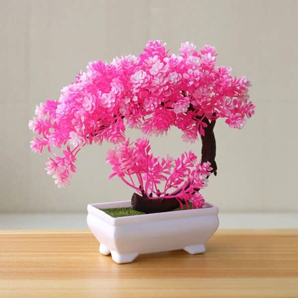 Un bonsaï artificiel de cerisier en fleurs rose vif dans un pot blanc sur une table en bois clair devant un mur blanc.